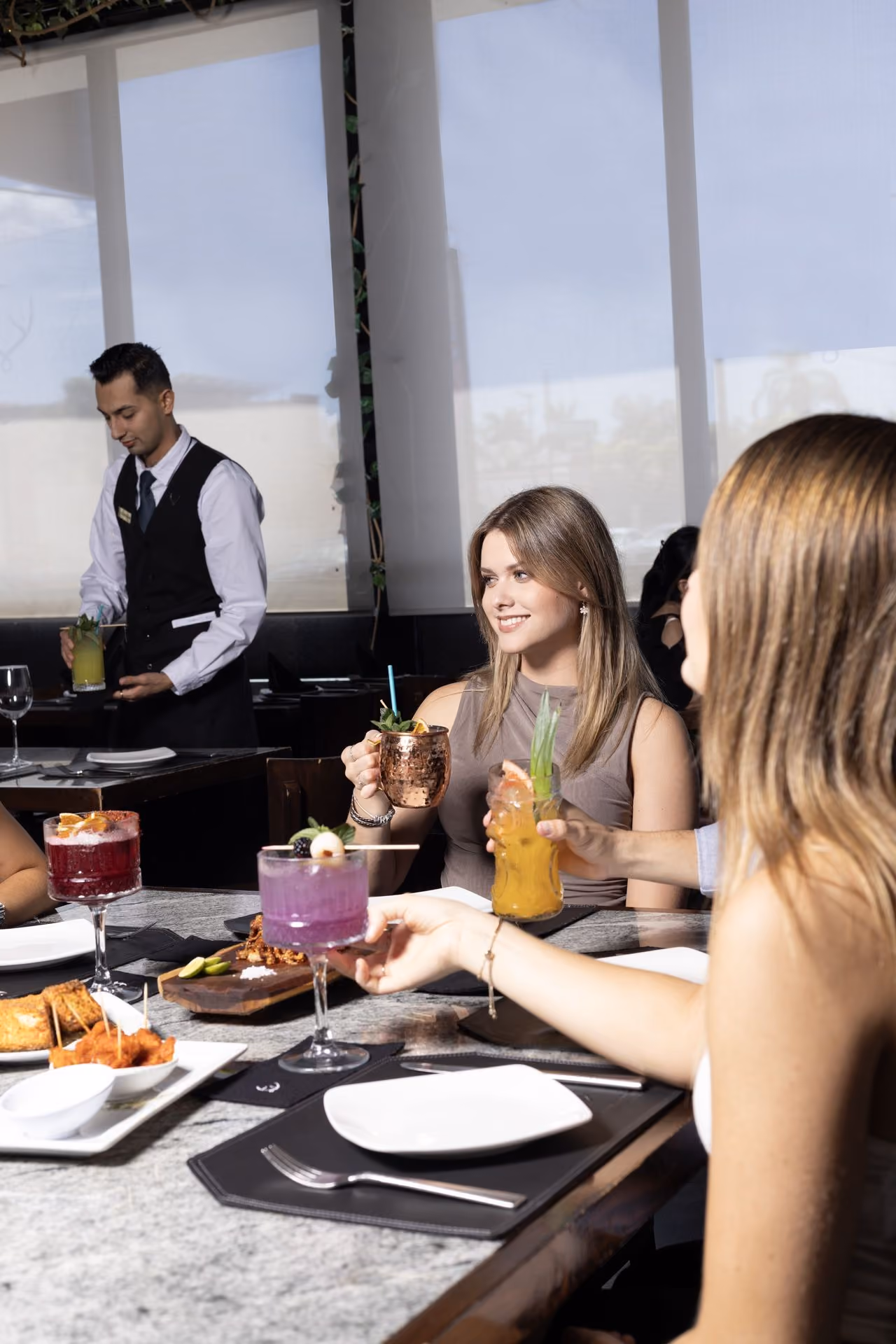 Amigas con cocteles junto al ventanal y mesero al fondo